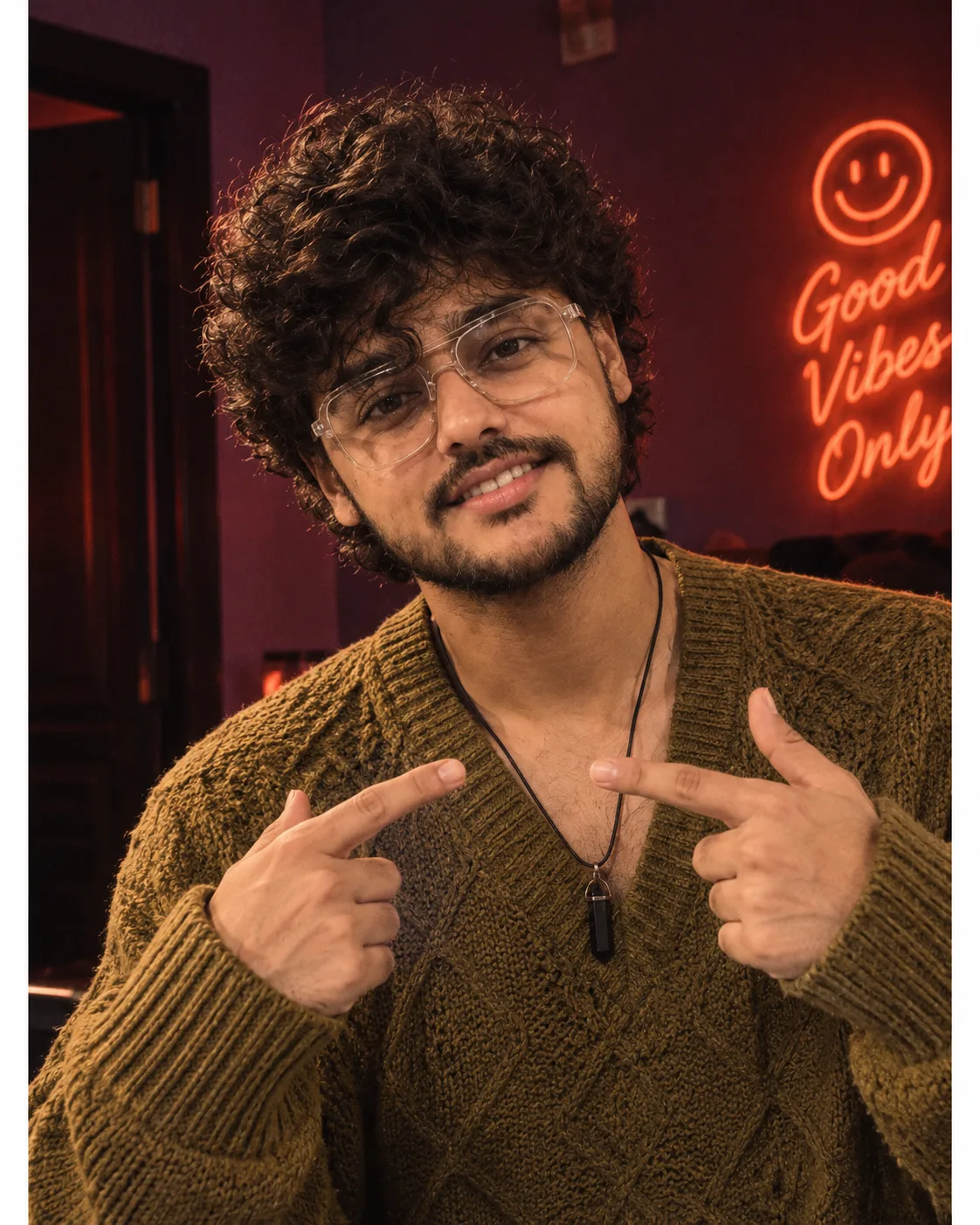 A playful young man wears an olive-mustard knit V-neck sweater.