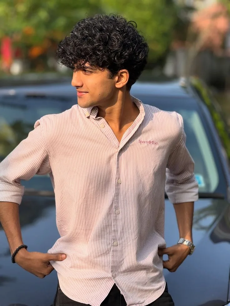 Candid Young Man in Pink Striped Shirt with Hands on Hips Beside Car in Afternoon Sunlight