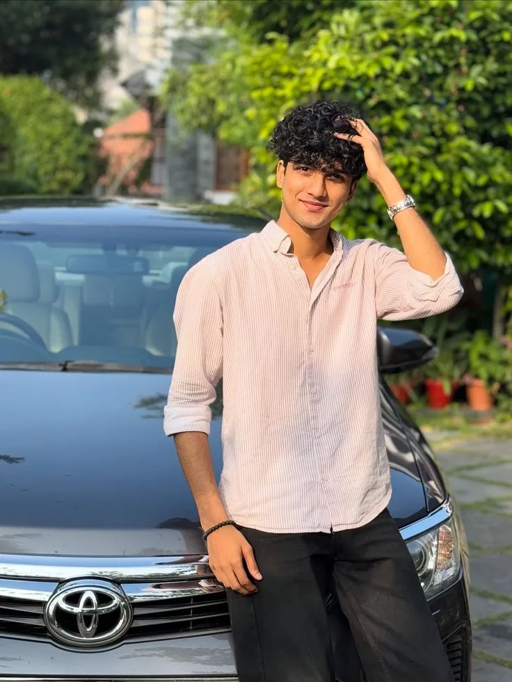 Candid Young Man in Pink Striped Shirt Running Hand Through Hair Beside Grey Car